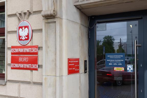 Drzwi wejściowe do budynku, obok których na ścianie wisi czerwona tablica z napisem "Biuro Rzecznika Praw Obywatelskich"