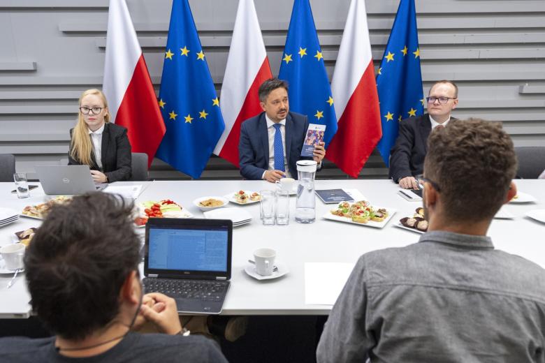 RPO Marcin Wiącek, dyrektor Piotr Mierzejewski, mec. Ewa Talma-Pogrzebska siedzący przy stole na tle flag Polski i UE
