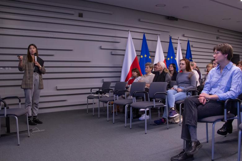 Prowadząca warsztat osoba wskazuje w kierunku prezentacji, a zgromadzona publiczność jej słucha. W tle flagi Polski i Unii Europejskiej