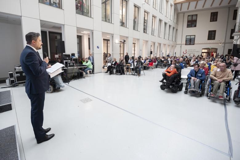 RPO Marcin Wiącek stoi i przemawia przed publicznością w dużej sali, pośród publiczności osoby na wózkach