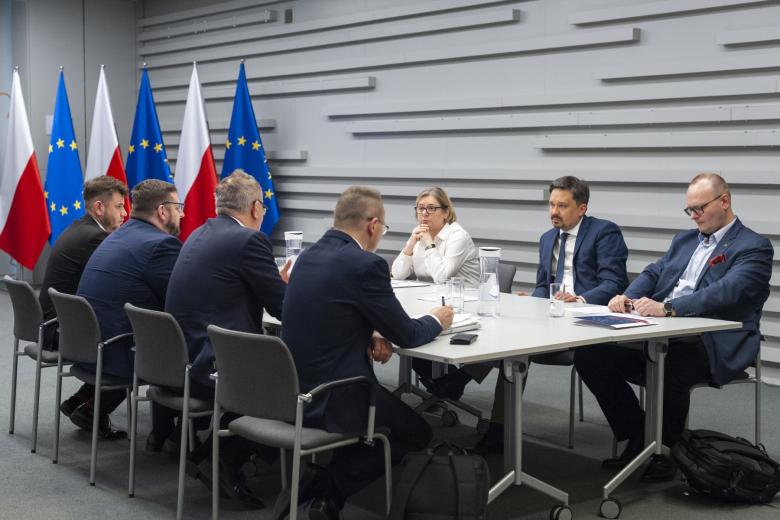 Siedem osób siedzi po obu stronach stołu i rozmawia. W tle flagi Polski i UE
