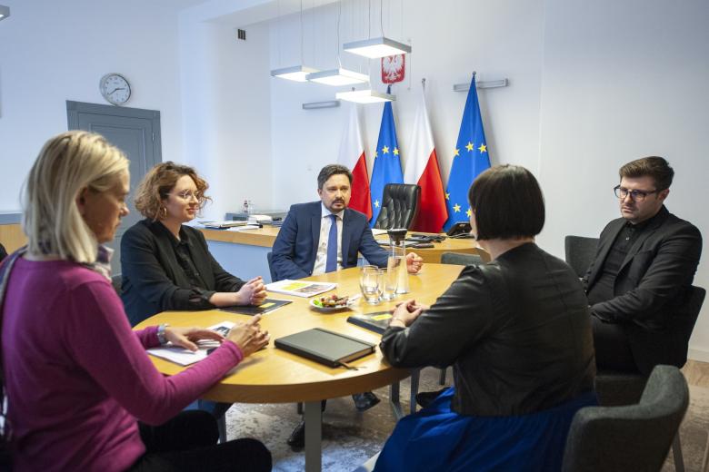 Pięć osób siedzi przy owalnym stole i rozmawia, w tle flagi Polski i UE