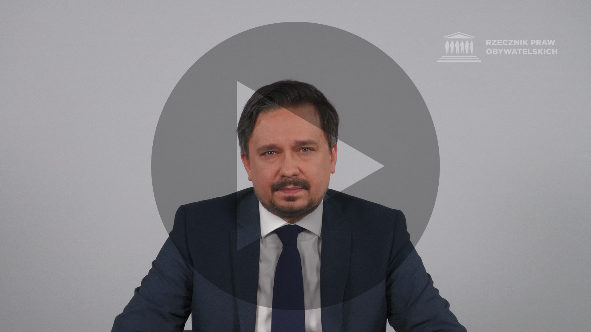 Kadr z nagrania przedstawiający RPO Marcina Wiącka z naniesionym symbolem odtwarzania wideo - trójkątem w kole