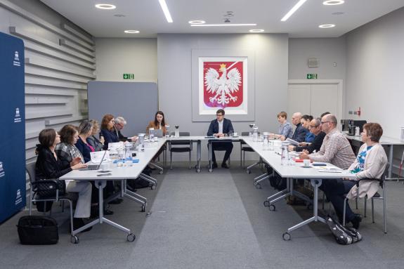 Kilkanaście osób siedzi przy stole konferencyjnym i rozmawia, w tle godło Polski z białym orłem na czerwonym tle