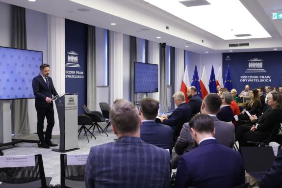 Kilkadziesiąt osób siedzi na sali konferencyjnej i słucha wystąpienia osoby mówiącej za mównicą na środku