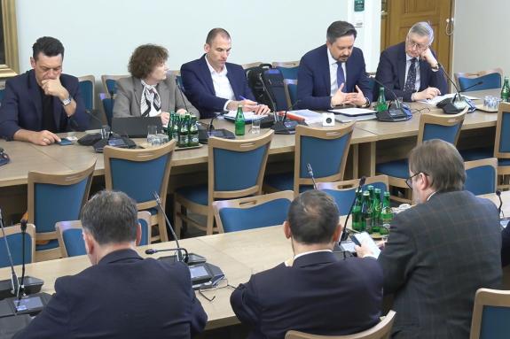 Kilkanaście osób siedzi przy stołach ustawionych w rzędy na dużej sali, na stołach stoja mikrofony