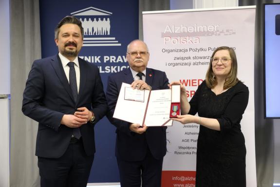 Trzy osoby pozują do wspólnego zdjęcia trzymając dyplom oraz odznakę w formie medalu