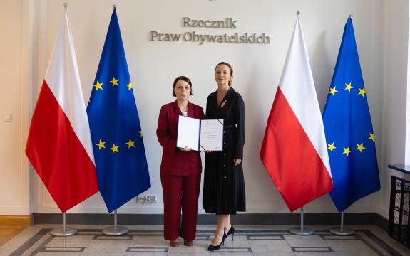 Sylwia Gregorczyk-Abram i Anna Błaszczak-Banasiak pozują do zdjęcia na tle flag Polski i Unii Europejskiej i napisu "Rzecznik Praw Obywatelskich", uśmiechając się. Anna Błaszczak-Banasiak trzyma w dłoniach otwartą teczkę z dokumentem powołania jej na stanowisko.