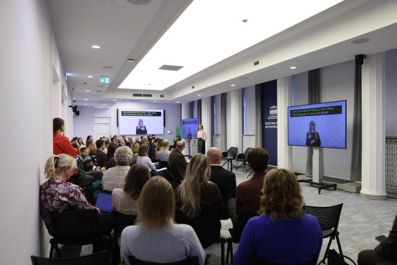 Widok ogólny sali konferencyjnej, na której siedzi kilkadziesiąt osób. Na środku przy mównicy stoi kobieta w beżowej bluzce i wypowiada się. Na wiszącym na ścianie ekranie wyświetla się tłumaczka języka migowego i napisy.