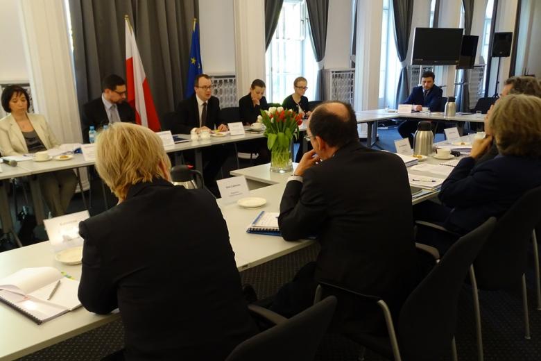 Zdjęcie: ludzie siedzą przy stole w formie kwadratu. Białe i czerwone tulipany, flaga polska i UE w tle