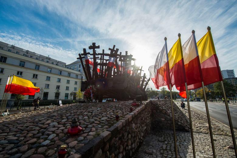 zdjęcie: pomnik przedstawiający wagon kolejowy z krzyżami, przed nim stoją flagi - Polski i Warszawy