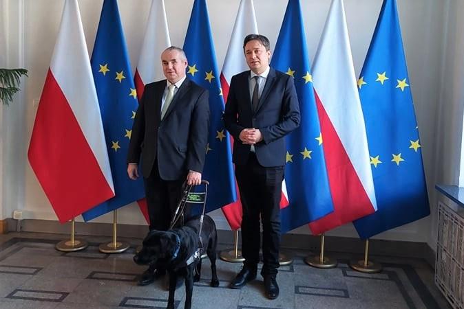 dwóch meżczyzn i pies przewodnik na tle flag polskich i UE