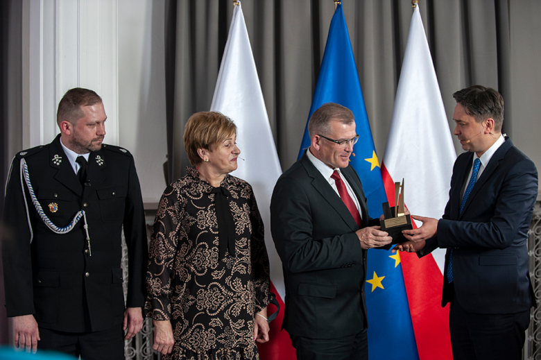 Mężczyzna wręcza nagrodę trzem laureatom, w tle flagi Polski i Unii Europejskiej