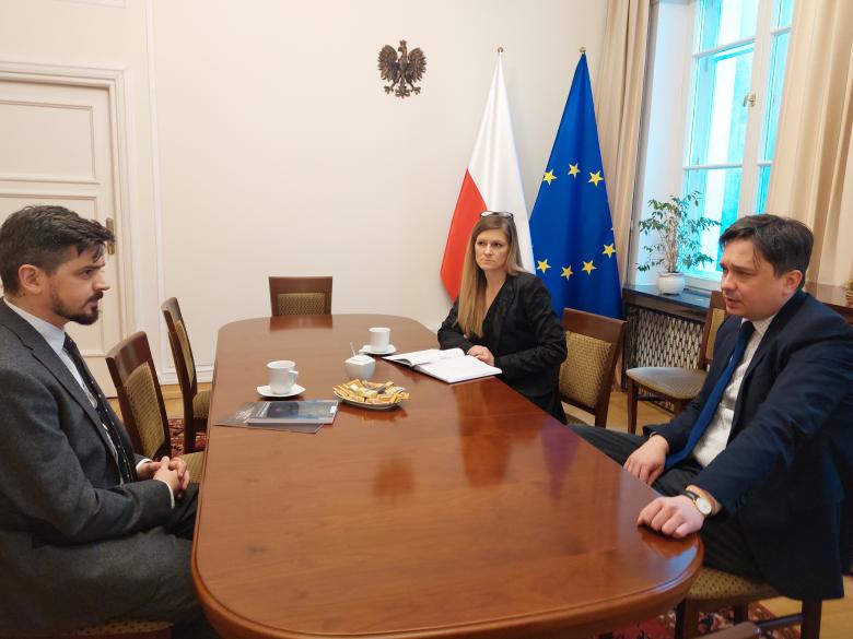Trzy osoby rozmawiające przy konferencyjnym stole. W rogu pokoju stoją flagi Polski i Unii Europejskiej, a na ścianie wisi godło państwowe.