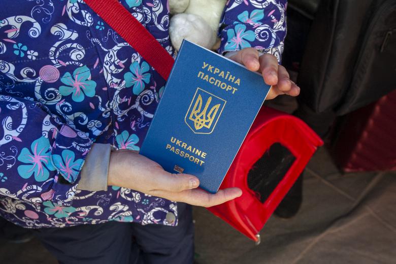 Zbliżenie na ukraiński paszport w dłoniach kobiety trzymającej pluszową zabawkę pod pachą oraz transporter dla kota przewieszony przez ramię.
