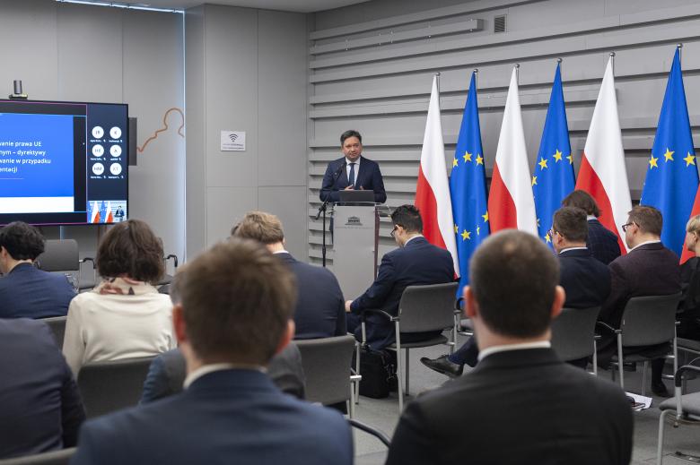 RPO Marcin Wiącek wypowiadający się przy podium w sali konferencyjnej. Przed RPO siedzą uczestnicy konferencji, a po jego prawej stronie znajduje się ekran przedstawiający wideokonferencję. Na lewo od RPO stoją flagi Polski i Unii Europejskiej