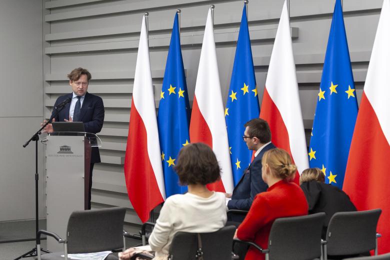 ZRPO Maciej Taborowski wypowiadający się z mównicy w stronę uczestników spotkania. Po lewej stronie ZRPO - flagi Polski i Unii Europejskiej
