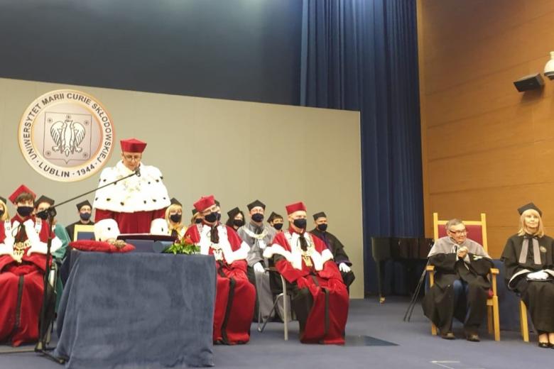 Rektor UMCS przemawia na uroczystości nadania tytułu doctora honoris causa Marianowi Turskiemu. Za rektorem siedzą przedstawiciele społeczności naukowej uniwersytetu, a po drugiej stronie, na honorowym miejscu, siedzi Marian Turski