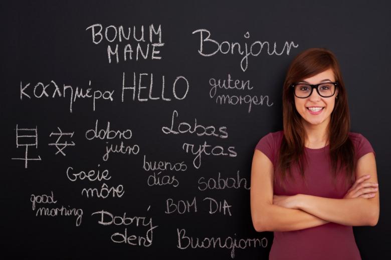 Osoba stojąca przed tablicą szkolną, na której kredą napisano powitanie w kilkunastu obcych językach