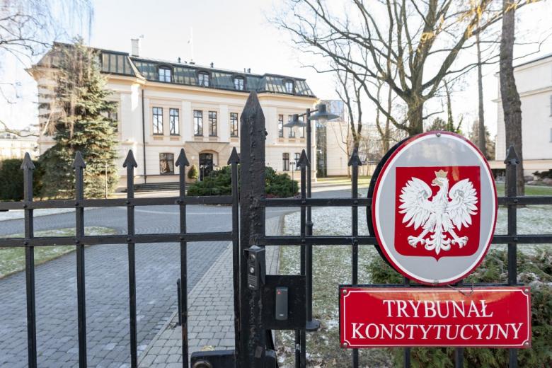 na pierwszym planie dwie tablice, jedna z godłem Polski, druga z napisem Trybunał Konstytucyjny, w oddali za ogrodzeniem budynek