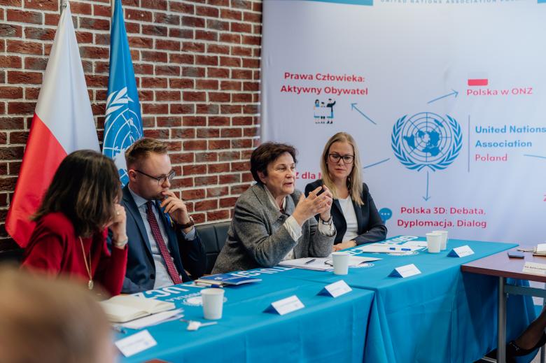 Cztery osoby, w tym ZRPO Hanna Machińska, siedzi w panelu przy długim stole. Wypowiada się Hanna Machińska, reszta osób słucha w skupieniu. W tle flagi Polski, ONZ oraz ścianka stowarzyszenia UNA Poland.