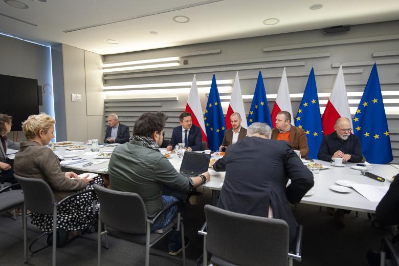RPO Marcin Wiącek rozmawiający z dziennikarzami przy konferencyjnym stole. W tle flagi Polski i Unii Europejskiej