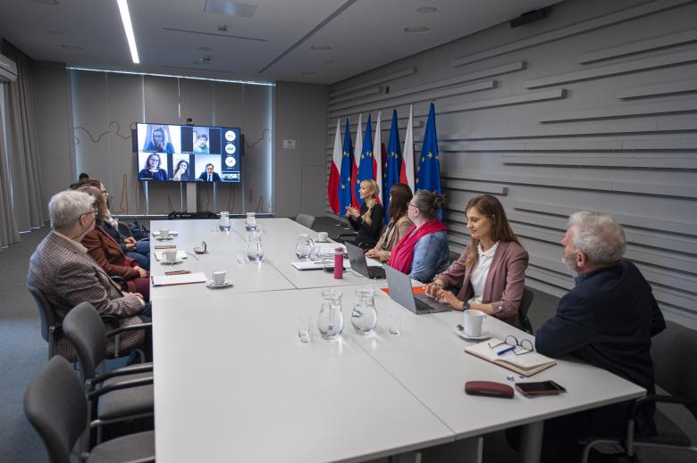 Szerokokątne ujęcie sali konferencyjnej, w której przy stole siedzą stacjonarni uczestnicy posiedzenia, a na ekranie konferencyjnym wyświetlone są sylwetki uczestników łączących się za pośrednictwem internetu. Pod ścianą stoją flagi Polski i Unii Europejskiej.