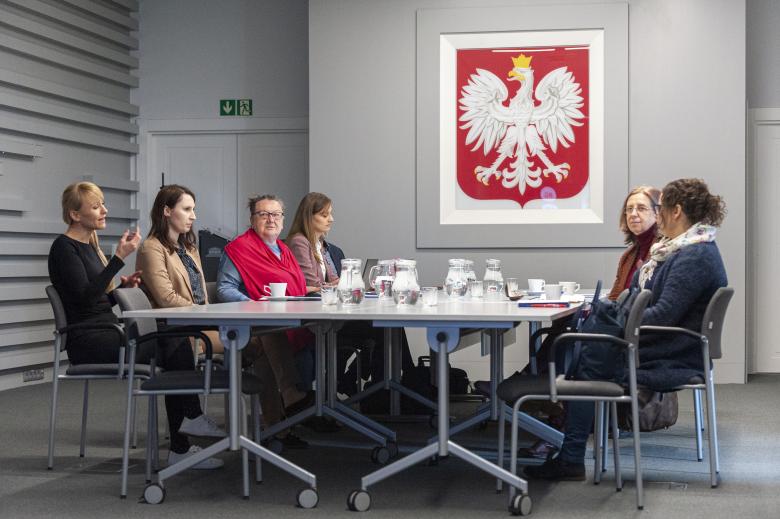 Stół konferencyjny, przy którym siedzą i dyskutują stacjonarni uczestnicy posiedzenia. Na ścianie u szczytu stołu znajduje się duże godło Polski.