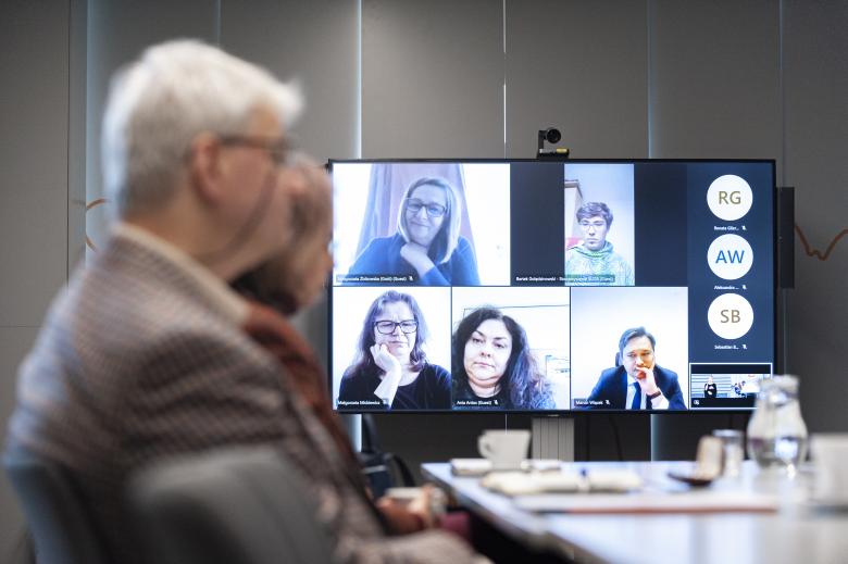 Zbliżenie na ekran pokazujący osoby uczestniczące w posiedzeniu za pośrednictwem internetu. Na pierwszym planie rozmyte sylwetki uczestników stacjonarnych.