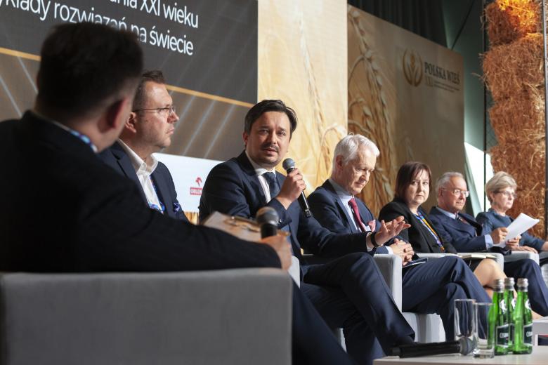 RPO Marcin Wiącek siedzi w rzędzie foteli razem z innymi panelistami i wypowiada się w stronę moderatora siedzącego w fotelu zwróconym w stronę panelistów. 