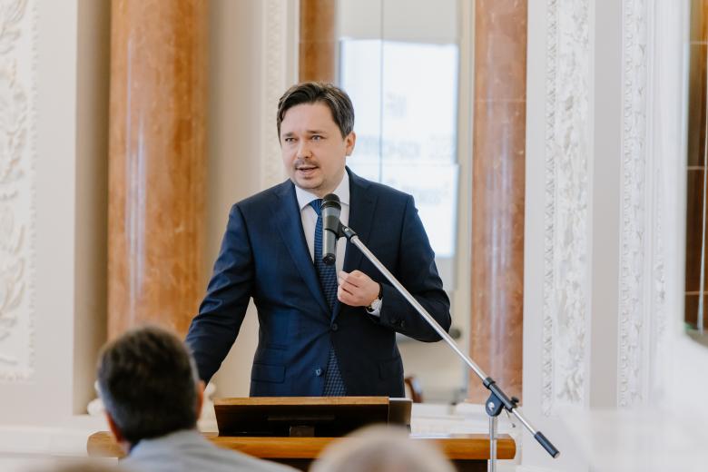 RPO Marcin Wiącek, stojący przy pulpicie przodem do słuchającej go widowni, wygłasza swój referat do mikrofonu w uniwersyteckiej sali. 
