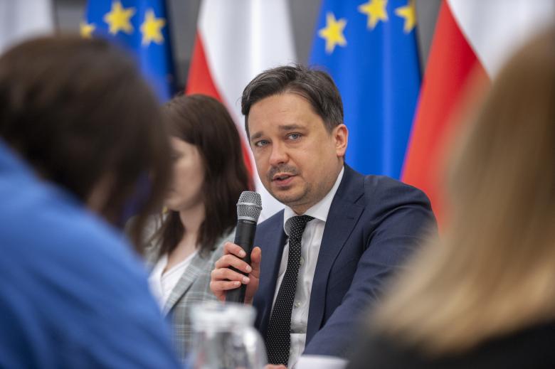 Mężczyzna mówi do mikrofonu, siedzi wśród ludzi, w tle flagi Polski i Unii Europejskiej