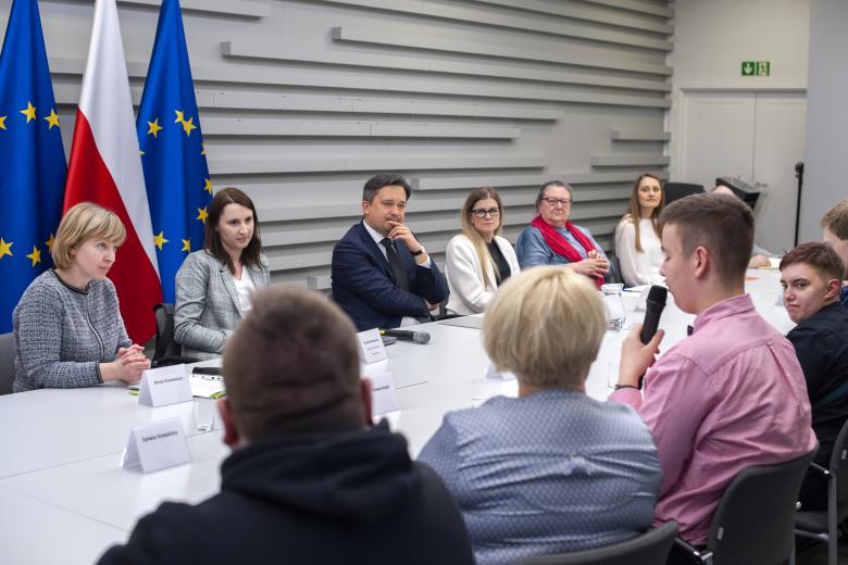 Ludzie siedzą przy długim stole, mężczyzna mówi do mikrofonu, pozostali słuchają, w tle flagi Polski i Unii Europejskiej