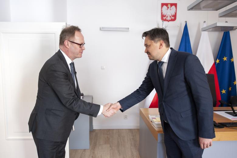 RPO Marcin Wiącek ściskający dłoń z ambasadorem Finlandii. W tle godło Polski oraz flagi Polski i Unii Europejskiej