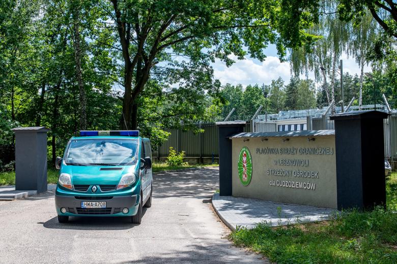 Zielony samochód Straży Granicznej wyjeżdzający przez bramę, na ogrodzeniu napis Placówka Straży Granicznej w Lesznowoli Strzeżony Ośrodek dla Cudzoziemców