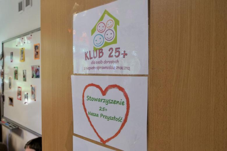 Zbliżenie na drzwi do klubu, na którym znajdują się wydrukowane loga Klubu 25+ i stowarzyszenia 25+ Nasza Przyszłość