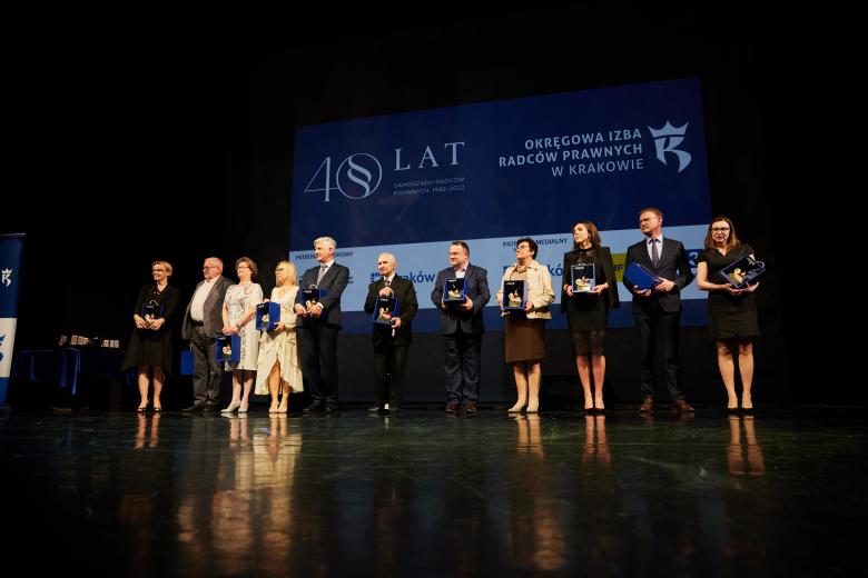 Uczestnicy gali, wśród nich ZRPO Hanna Machińska, stoją na scenie trzymając okolicznościowe podarunki - skrzynki z figurkami przedstawiającymi krakowskiego lajkonika
