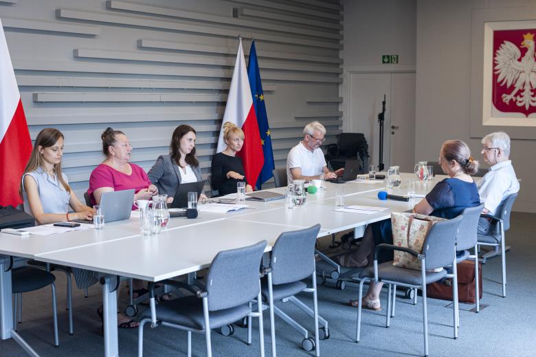 Członkowie komisji ekspertów rozmawiający przy konferencyjnym stole. Jedna z siedzących osób tłumaczy rozmowę na język migowy. W tle godło Polski oraz flagi Polski i Unii Europejskiej