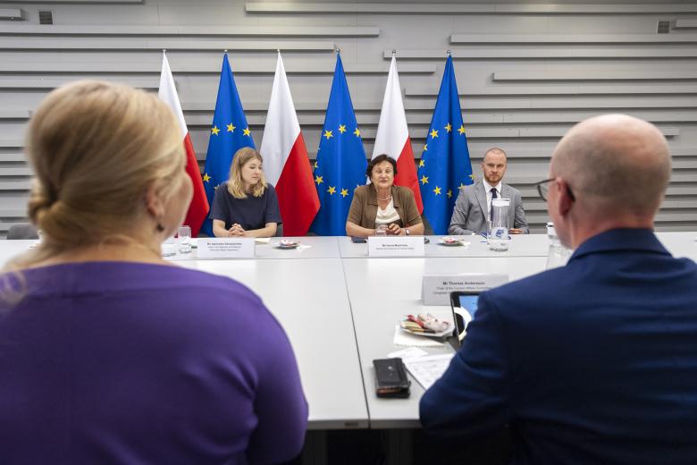 ZRPO Hanna Machińska, siedząca razem z pracownikami biura RPO, wypowiada się w stronę siedzących tyłem do kadru gości. W tle flagi Polski i Unii Europejskiej