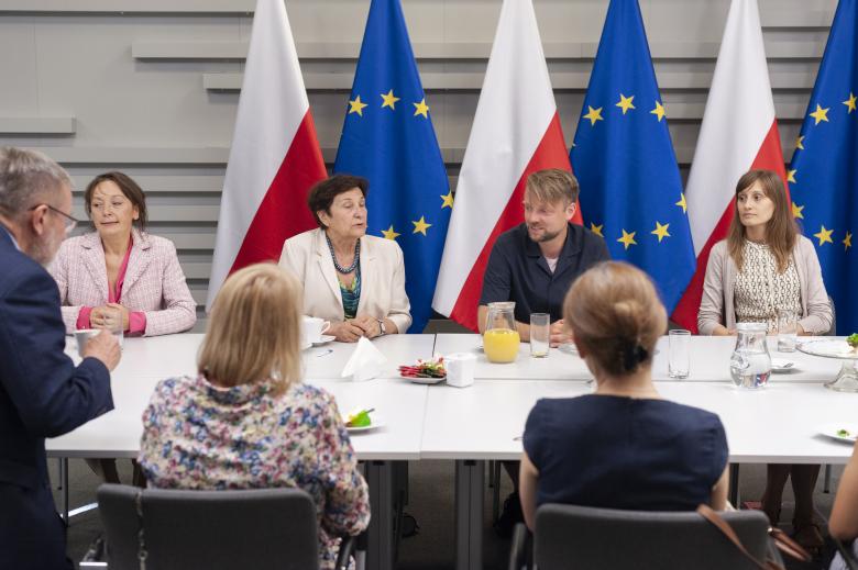 Grupa ludzi siedzi przy prostokątnym stole, w tle flagi Polski i Unii Europejskiej