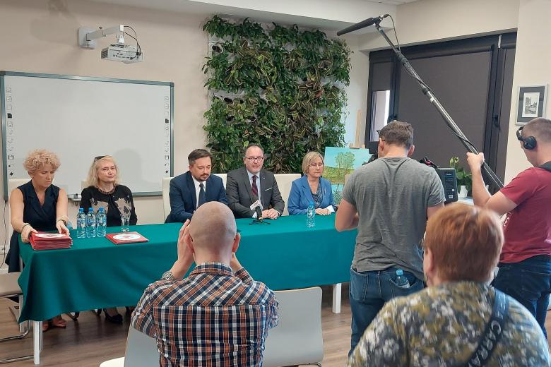 5 osób siedzi za stołem a dziennikarze zadają pytania