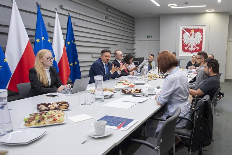 Grupa osób siedzących przy stole konferencyjnym, w tle flagi Polski i UE