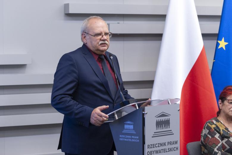 Prezes Związku Stowarzyszeń Alzheimer Polska Zygmunt Wierzyński przemawia za mównicą, w tle flaga Polski i UE