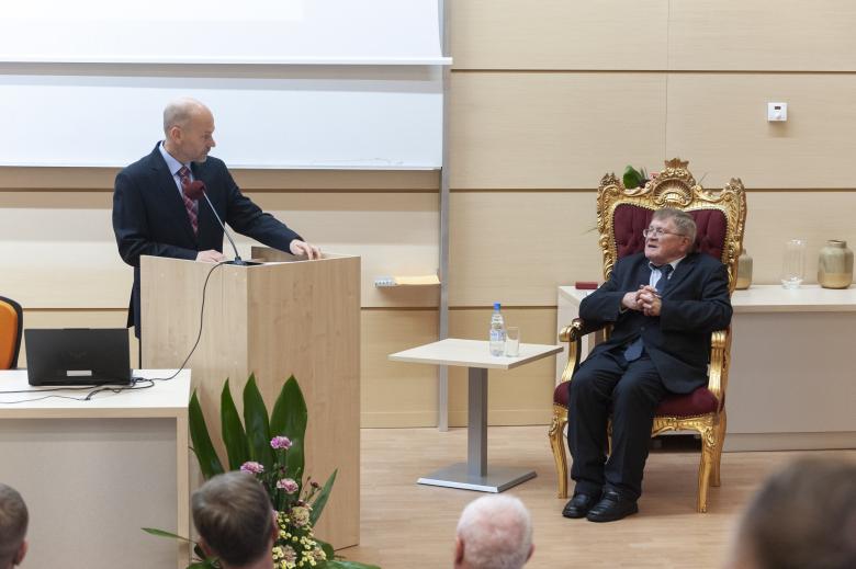 Dyrektor Generalny BRPO Maciej Berek wypowiada się do mikrofonu stojąc przy podium, patrząc na siedzącego w fotelu profesora Grzybowskiego