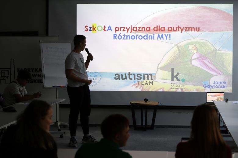 Uczestnik spotkania wypowiada się do mikrofonu przedstawiając wyświetlaną na ścianie prezentację pod tytułem "Szkoła przyjazna dla autyzmu - różnorodni my!"