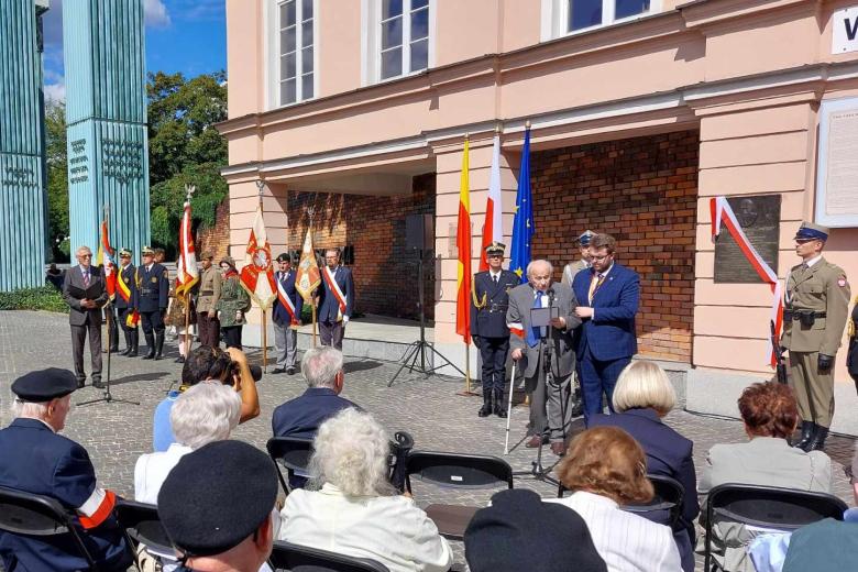 Powstaniec warszawski wypowiada się do mikrofonu, stojąc przed tablicą pamiątkową. Obok tablicy stoją żołnierze pełniący wartę.