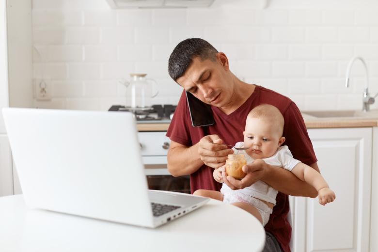 Mężczyzna obrany w bordową koszulkę karmi łyżeczką małe dziecko siedzące mu na kolanach przy stole w kuchni i rozmawiający przez telefon. Na stole przed nim otwarty laptop.