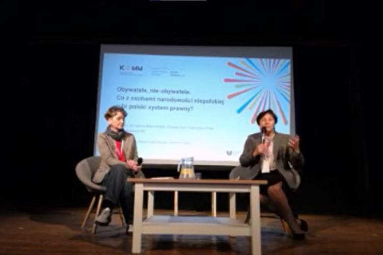 Dwie osoby siedzą w fotelach przy stole na scenie, w tle ekran z napisem "Forum Partnerstwa i Integracji. Obywatele nie-obywatele. Co z osobami narodowości niepolskiej robi polski system prawny"