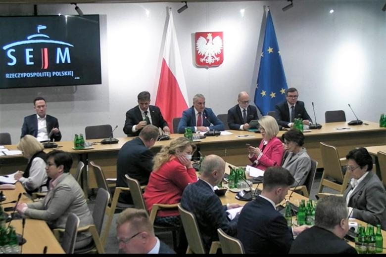 Kilkadziesiąt osób siedzi na sali przy stołach, na ścianie godło Polski, obok stoją flagi Polski i UE, na ścianie ekran z napisem "SEJM Rzeczypospolitej Polskiej"