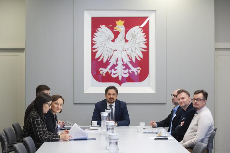 RPO Marcin Wiącek i kilkoro osób siedzących przy stole konferencyjnym, w tle godło Polski